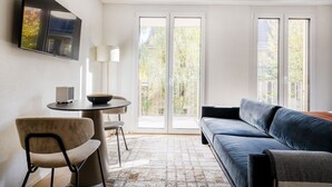 Un salon moderne comprenant un canapé, une table à manger avec des chaises, un téléviseur fixé au mur et de grandes fenêtres donnant sur un paysage verdoyant.