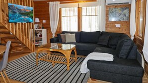 Un intérieur de chalet chaleureux avec un canapé d’angle, une table basse en verre et un téléviseur fixé à un mur en bois.