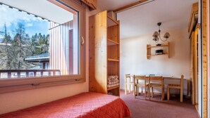 A bedroom with a bed, a desk, and a chair, a window with a view of trees, and a wooden shelf. - Studio 4 ppl near the ski runs, ski lifts at 50m (Doucy)