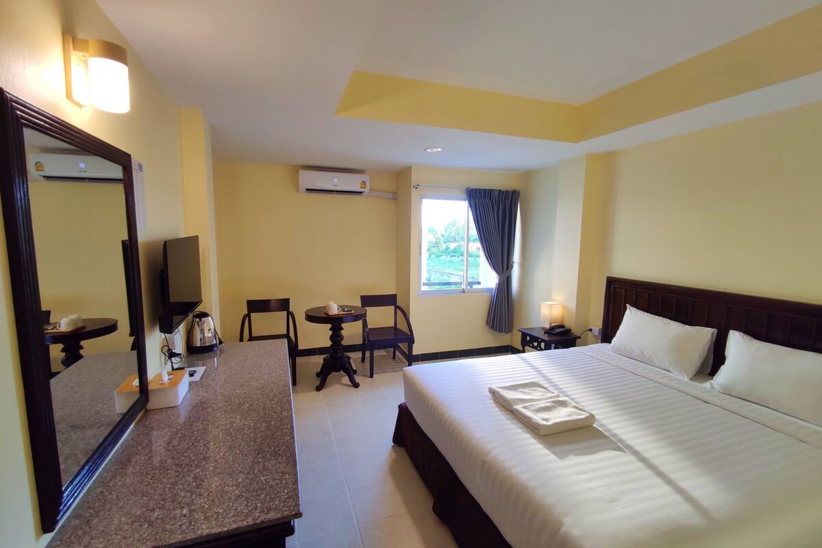Une chambre d’hôtel équipée d’un lit, d’un bureau, d’une chaise, d’une petite table, d’un miroir, d’un appareil de climatisation et d’une fenêtre avec des rideaux.