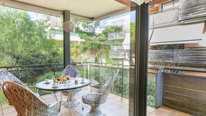 A balcony with a round table and wicker chairs, overlooking a garden and buildings.