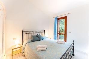 A bedroom with a bed, a nightstand, a window with curtains, and a view of greenery outside.
