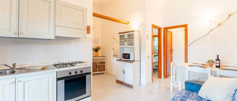 A compact kitchen with white cabinets, a stove, oven, and a microwave. There is a dining area with a table and chairs, and a bed with a blue and white patterned bedspread.