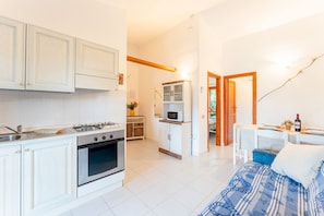 A compact kitchen with white cabinets, a stove, oven, and a microwave. There is a dining area with a table and chairs, and a bed with a blue and white patterned bedspread. - Residenze Gallura, three-room apartment 4 beds with air conditioning (San Teodoro)