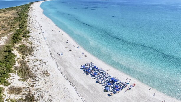 Oversigtsbillede af en strand med klart turkis vand, en sandet kystlinje og et bjergrigt bagland.