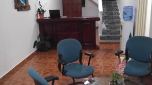 Uma sala de conferências com uma mesa de vidro, quatro cadeiras azuis, uma escrivaninha de madeira, um laptop e uma escada ao fundo.