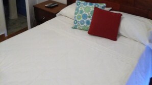 Quarto com cabeceira de madeira, cama com roupa de cama branca, mesa de cabeceira com um controle remoto e banheiro visível por uma porta aberta.