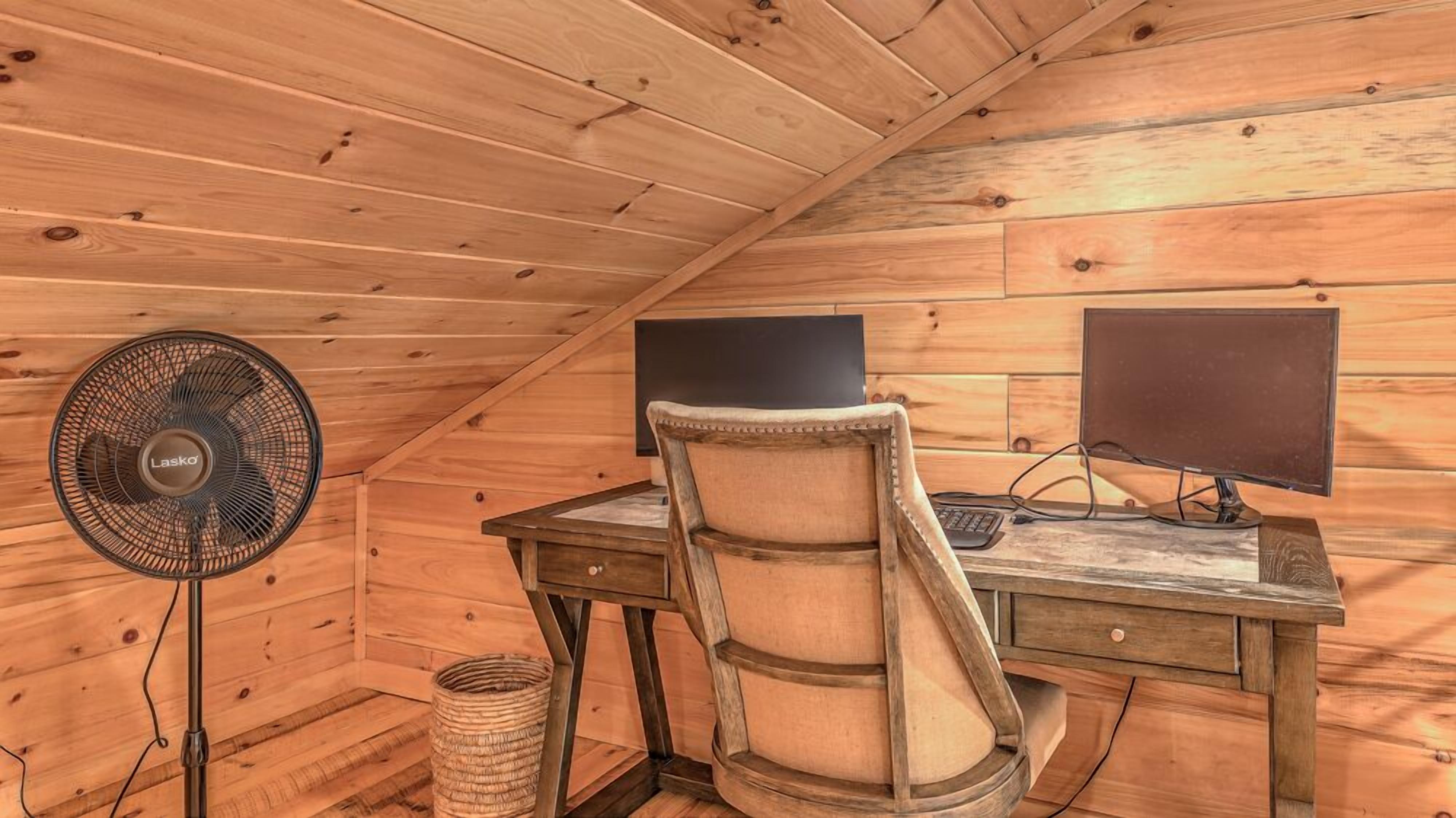 A wooden-paneled room with a desk, two monitors, a chair, and a standing fan.