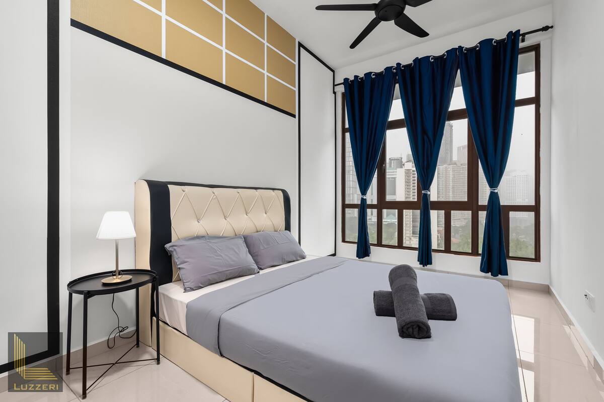 Une chambre moderne avec un lit, une table de chevet dotée d’une lampe, et une vue sur la ville grâce à de grandes fenêtres.