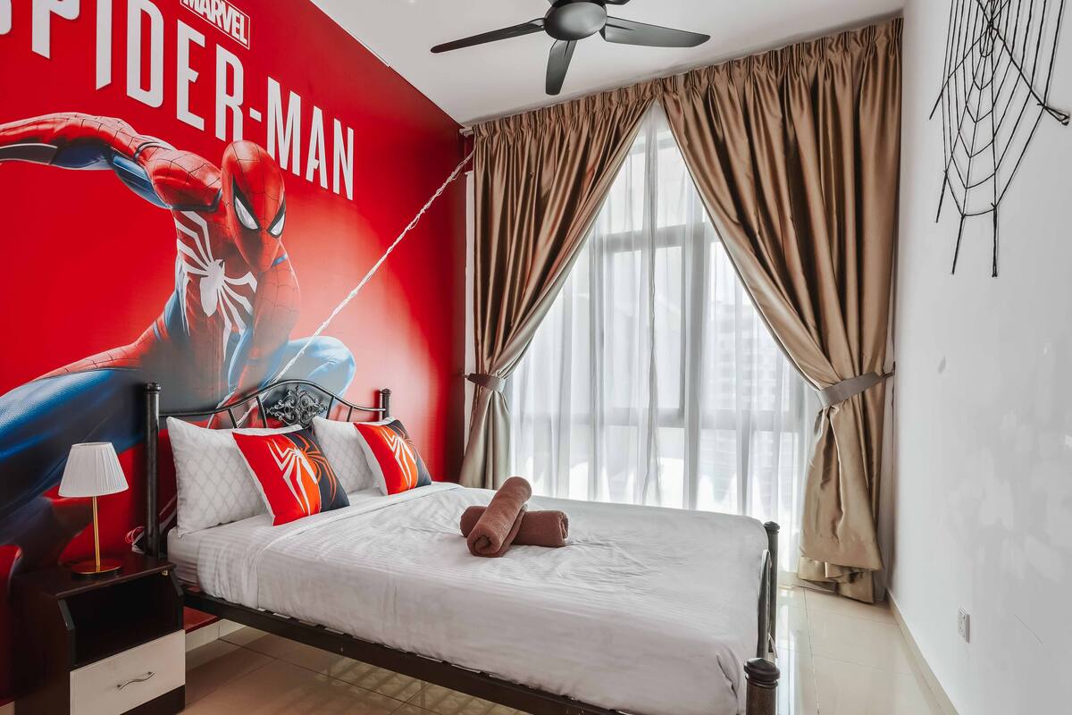 Une chambre avec une grande fresque représentant Spider-Man, un lit avec une literie blanche, une table de chevet, une lampe et un ventilateur de plafond.
