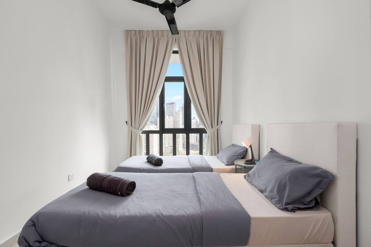 Une chambre d’hôtel moderne avec deux lits, une grande fenêtre avec des rideaux, un ventilateur de plafond et une vue sur le paysage urbain.