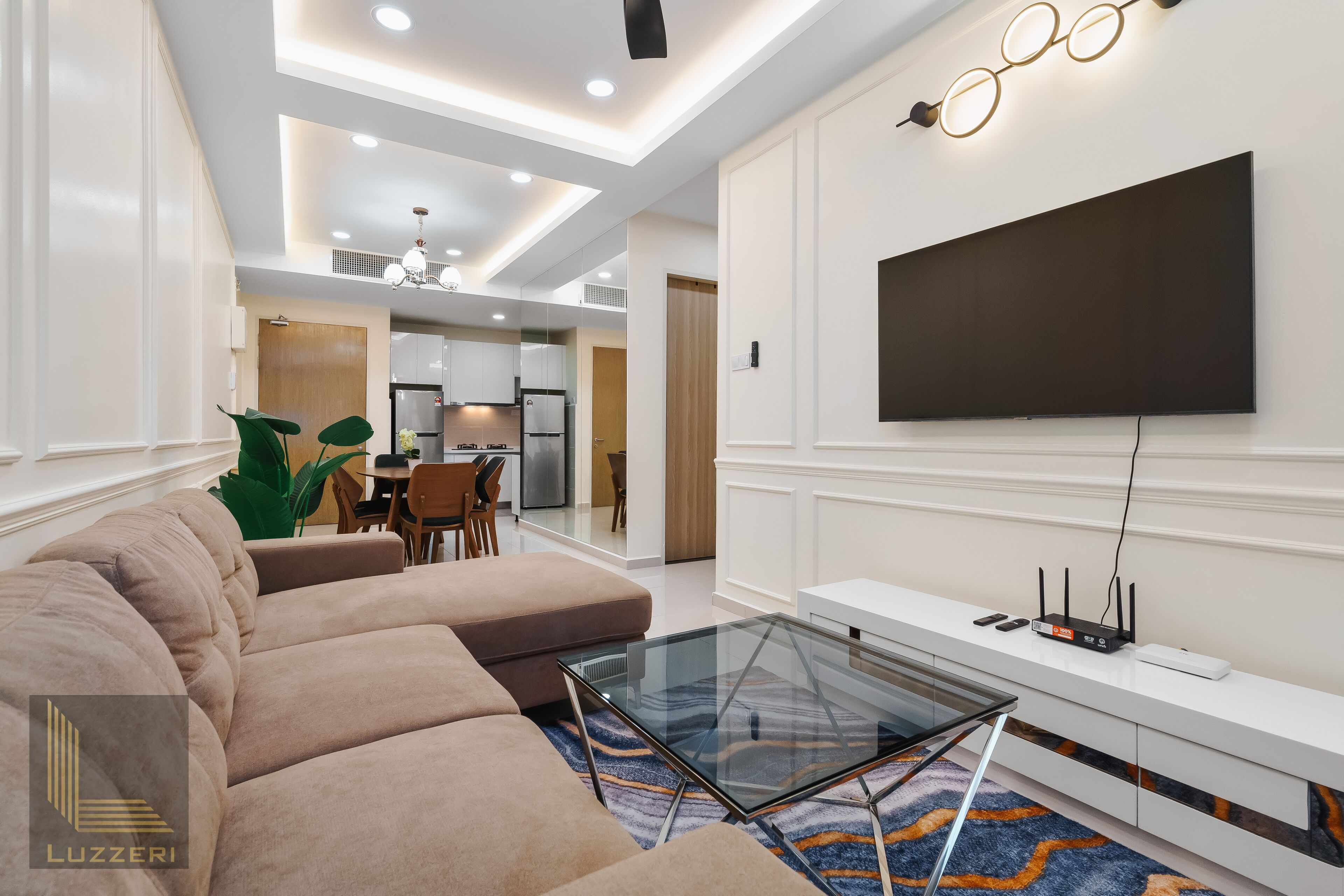 A modern living room with a large sectional sofa, a glass coffee table, a flat-screen TV mounted on the wall, and a dining area in the background.