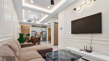 A modern living room with a large sectional sofa, a glass coffee table, a flat-screen TV mounted on the wall, and a dining area in the background.