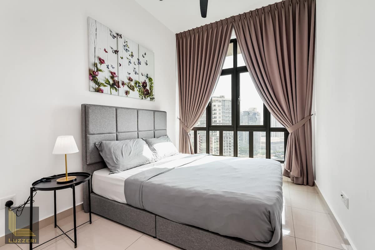 Une chambre moderne avec un lit gris, une table de chevet avec une lampe, une vue sur la ville à travers de grandes fenêtres et une œuvre d’art florale accrochée au mur.