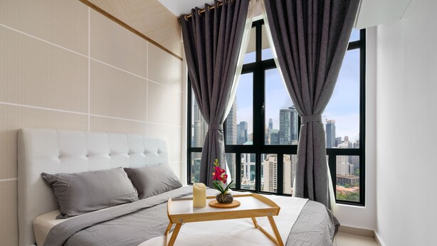 A modern bedroom with a large bed, a bedside table with a candle and flowers, and a city view through large windows. - Legasi Kampung Bharu Guesthouse (Kuala Lumpur)