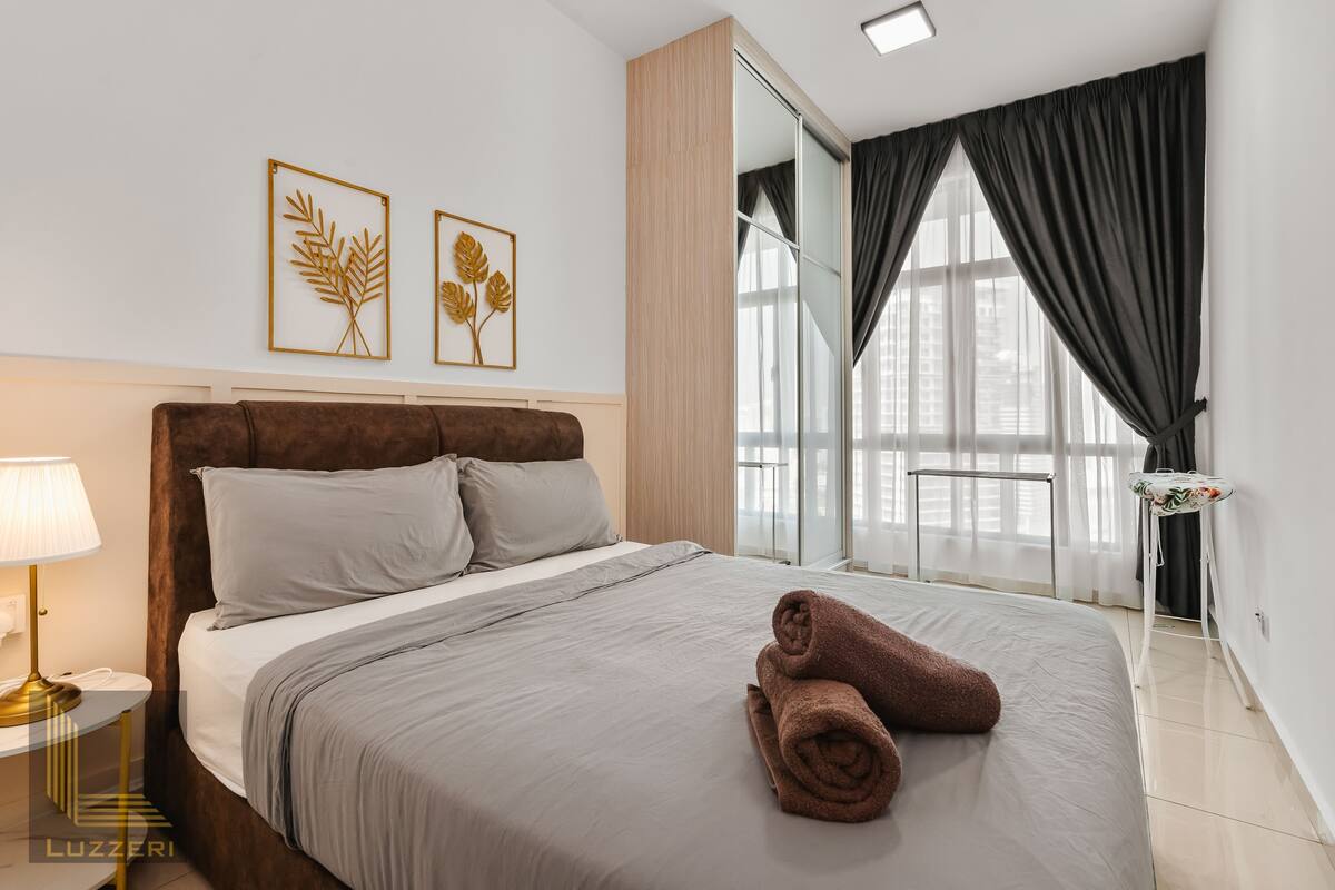 Un lit bien fait, avec une couvre-lit grise et deux oreillers, une tête de lit marron, une lampe de chevet et deux œuvres d’art représentant des feuilles, encadrées et accrochées au mur.
