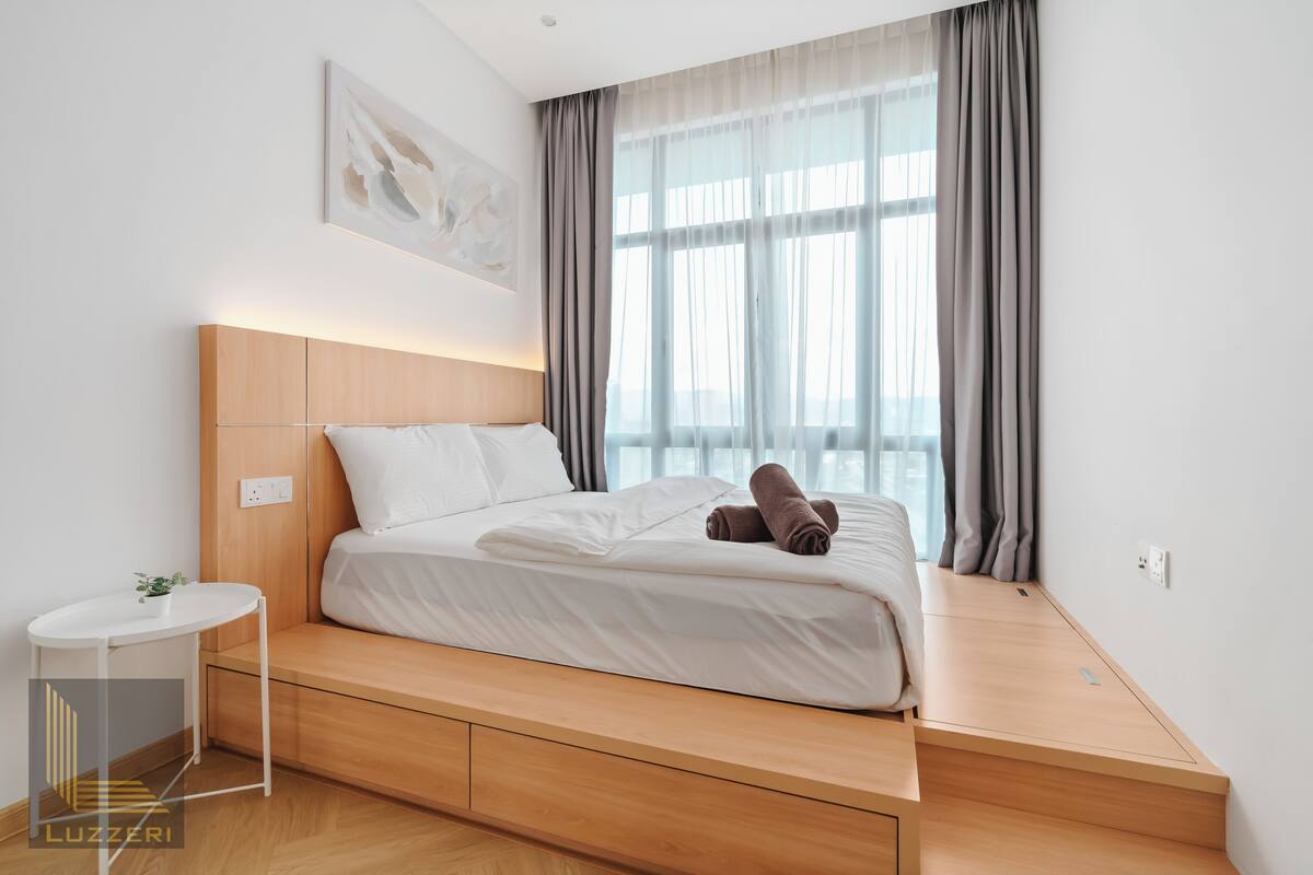 Une chambre d’hôtel moderne avec un lit sur une plateforme en bois, une petite table de chevet et une grande fenêtre avec des rideaux.