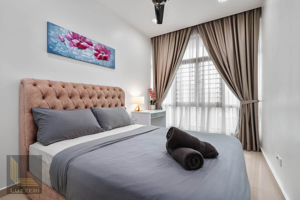 Une chambre à coucher avec une tête de lit capitonnée, un lit avec une literie grise, une serviette roulée, une table de chevet avec une lampe et une fenêtre avec des rideaux.