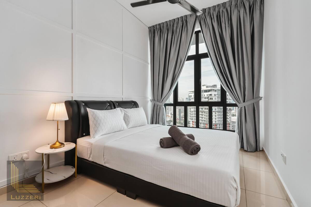 Une chambre d’hôtel moderne avec un grand lit, une table de chevet avec une lampe et une vue sur la ville grâce à de grandes fenêtres.