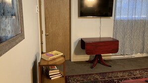 A living room with a wooden door, a flat-screen TV mounted on the wall, a wooden side table with books, and a patterned rug. - Cozy Newmarket Escape! Wine & WiFi! Full Kitchen! (Newmarket)