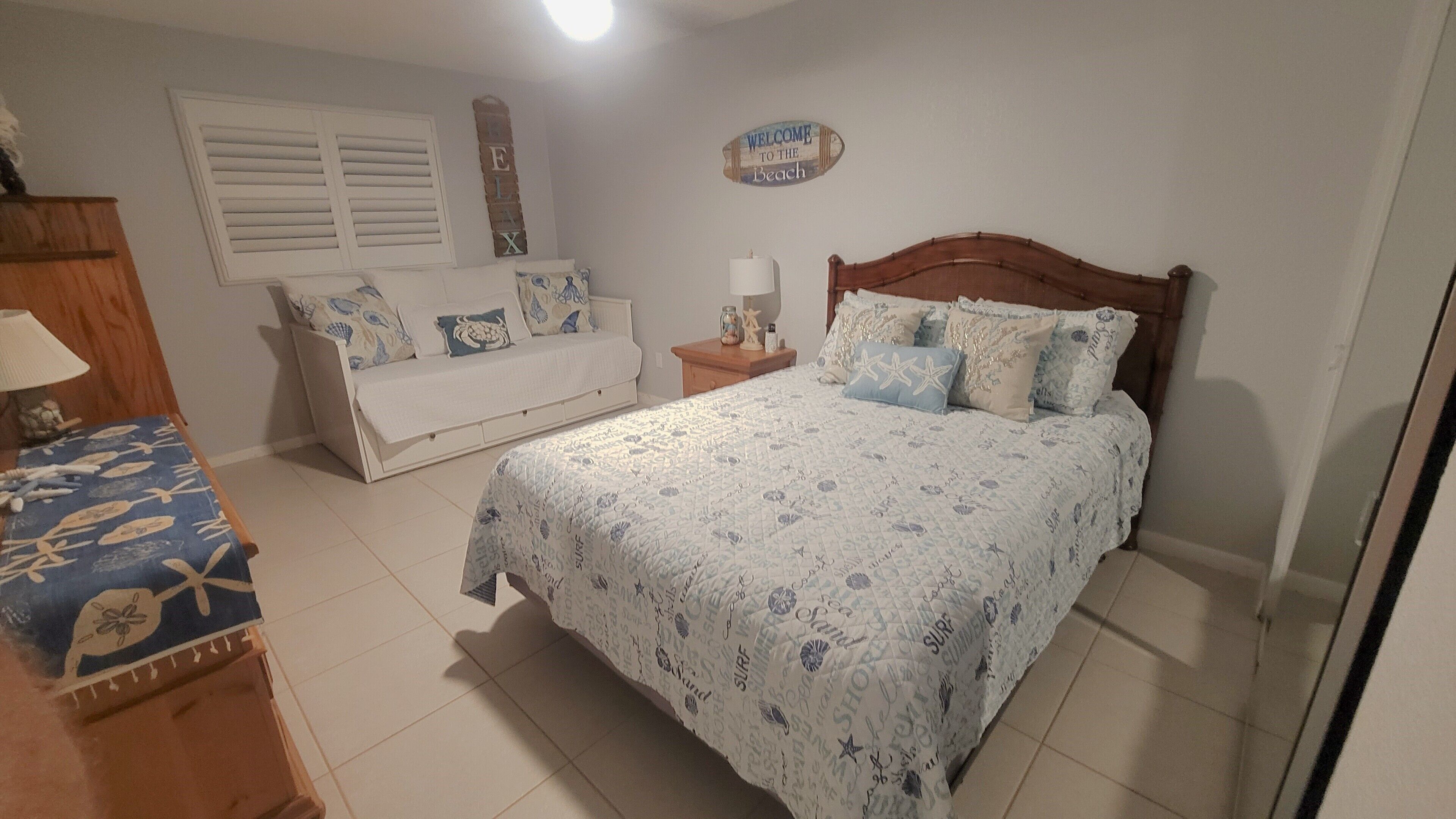 A bedroom with a bed, a nightstand, a dresser, a lamp, a window with shutters, and a wall sign.