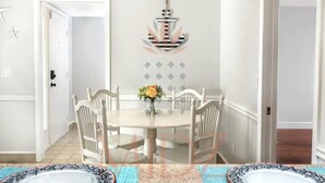 Un comedor con una mesa redonda, sillas blancas y un florero con flores. Se observa un ancla decorativa en la pared.