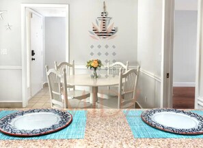 Un coin repas avec une table ronde, des chaises blanches et un vase rempli de fleurs. Un motif décoratif en forme d’ancre orne le mur.