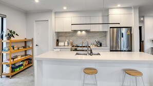 Uma cozinha moderna com armários brancos, uma geladeira de aço inoxidável, forno embutido e painel de fundo de mármore. Há dois bancos de madeira na ilha.