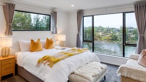 Quarto com uma cama grande, duas mesas de cabeceira, uma cadeira e vista para um rio e casas através de janelas amplas.