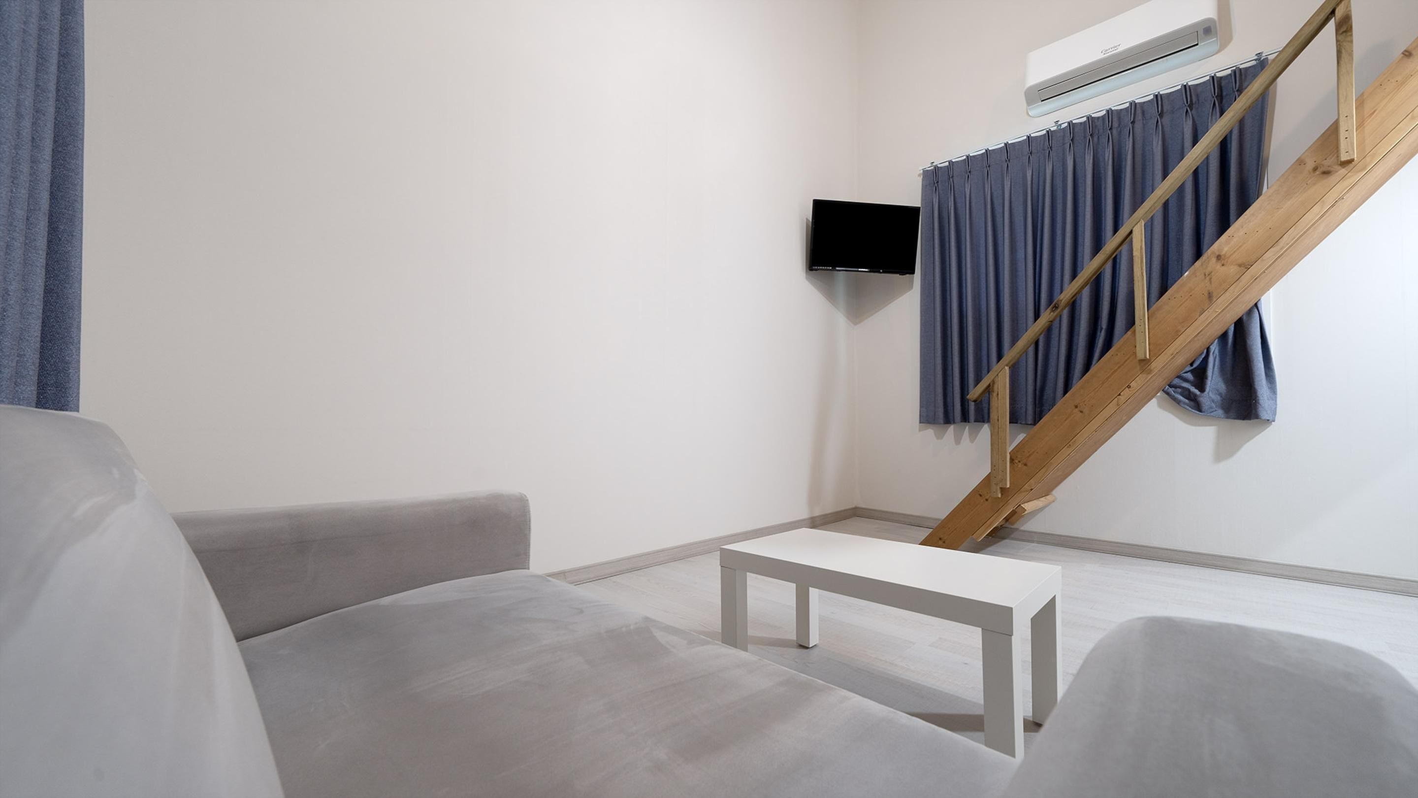 A modern, minimalist room with a grey sofa, a small white table, and a wooden staircase with blue curtains.