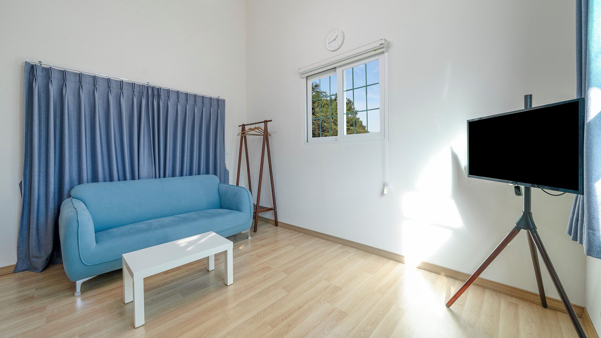 Une pièce avec un canapé bleu, un banc blanc, une fenêtre donnant sur un beau paysage et un téléviseur posé sur un trépied.