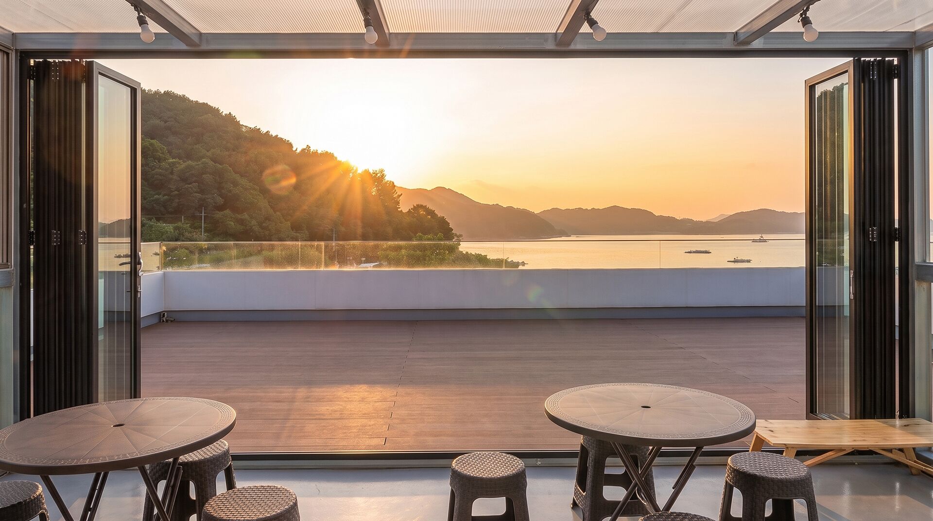 A modern outdoor terrace with glass doors, offering a view of a sunset over a body of water and hills.