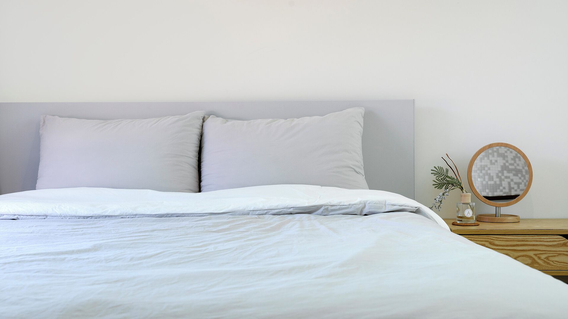 Una cama bien hecha con ropa blanca y almohadas, un espejo redondo con marco de madera sobre una mesita de noche, y un pequeño florero con una planta.