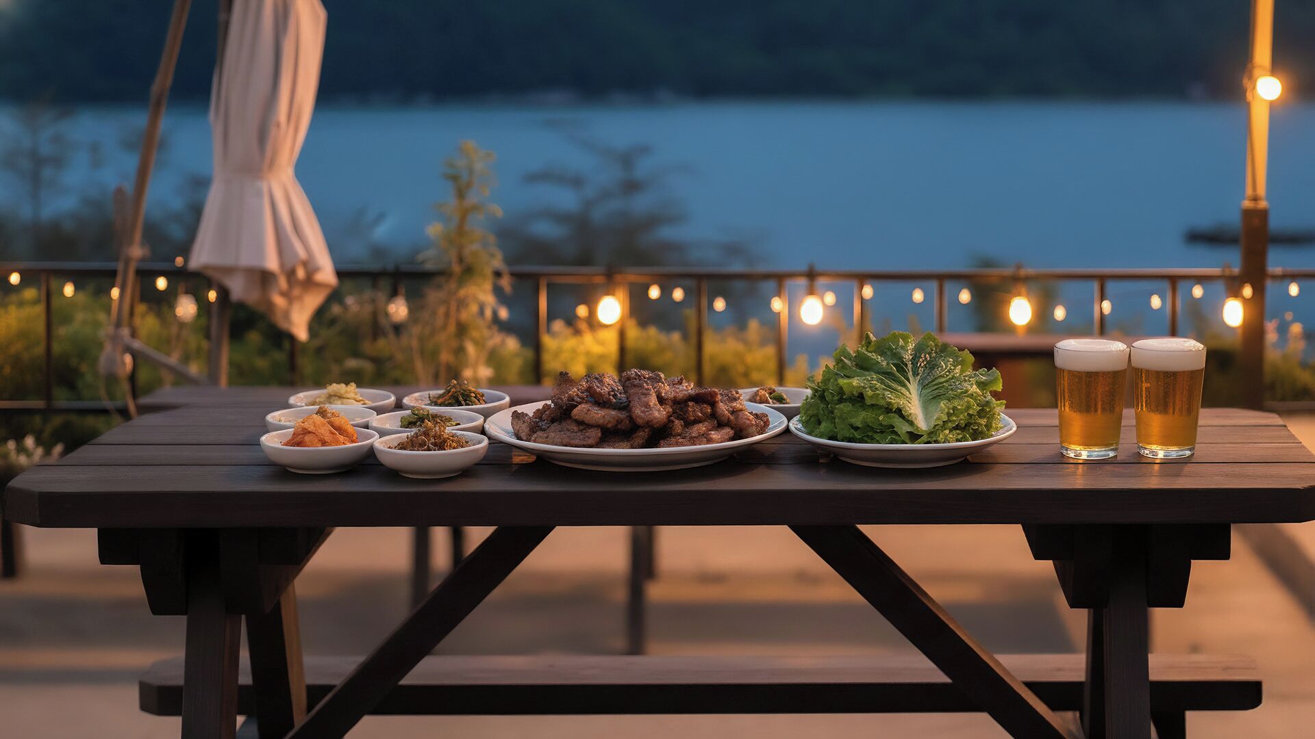 Una mesa de madera dispuesta con varios platos, una ensalada y dos vasos de cerveza, con vista a un cuerpo de agua.