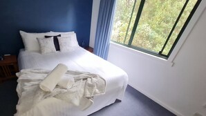 Un lit bien fait, avec du linge de lit blanc et une serviette pliée, une table de chevet avec une lampe et une fenêtre donnant sur des arbres.