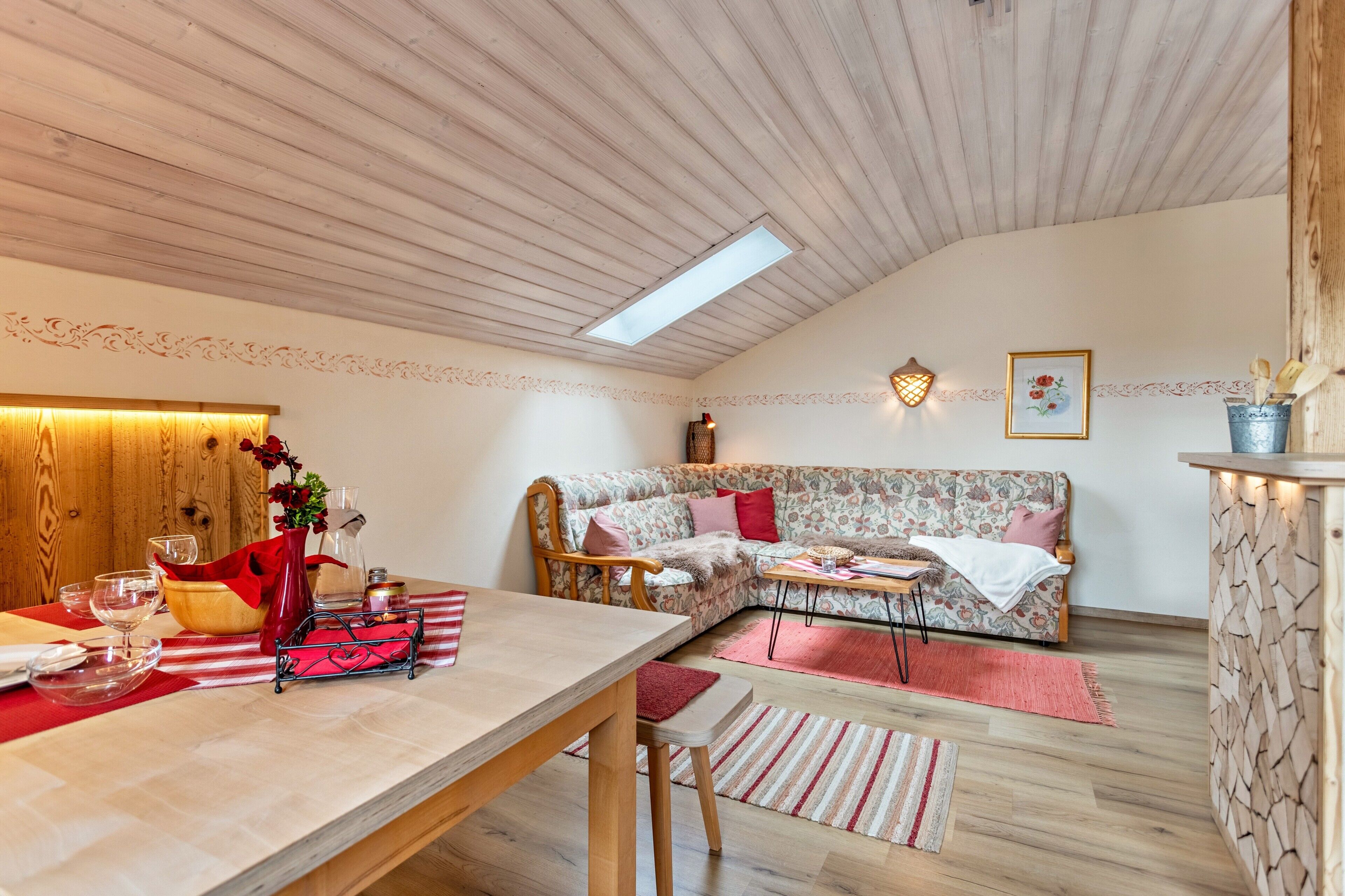Una stanza accogliente con soffitto in legno, un divano, un tavolo da pranzo con sedie, un angolo cottura e una parete decorativa.
