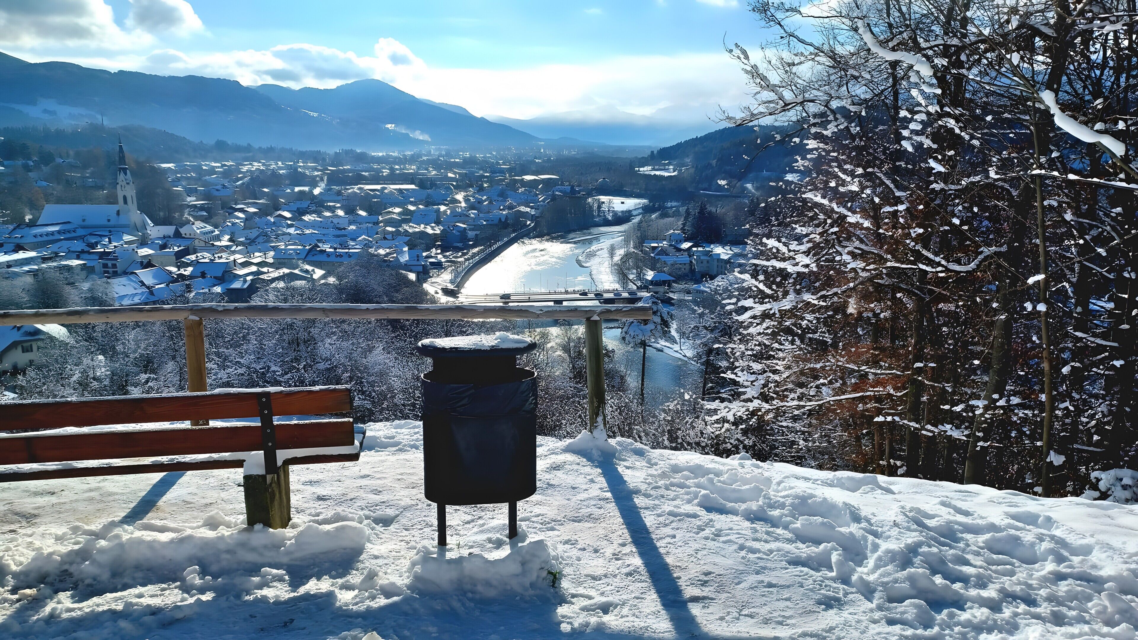 Un banc et une poubelle dans un décor extérieur enneigé, avec vue sur une ville et des montagnes.