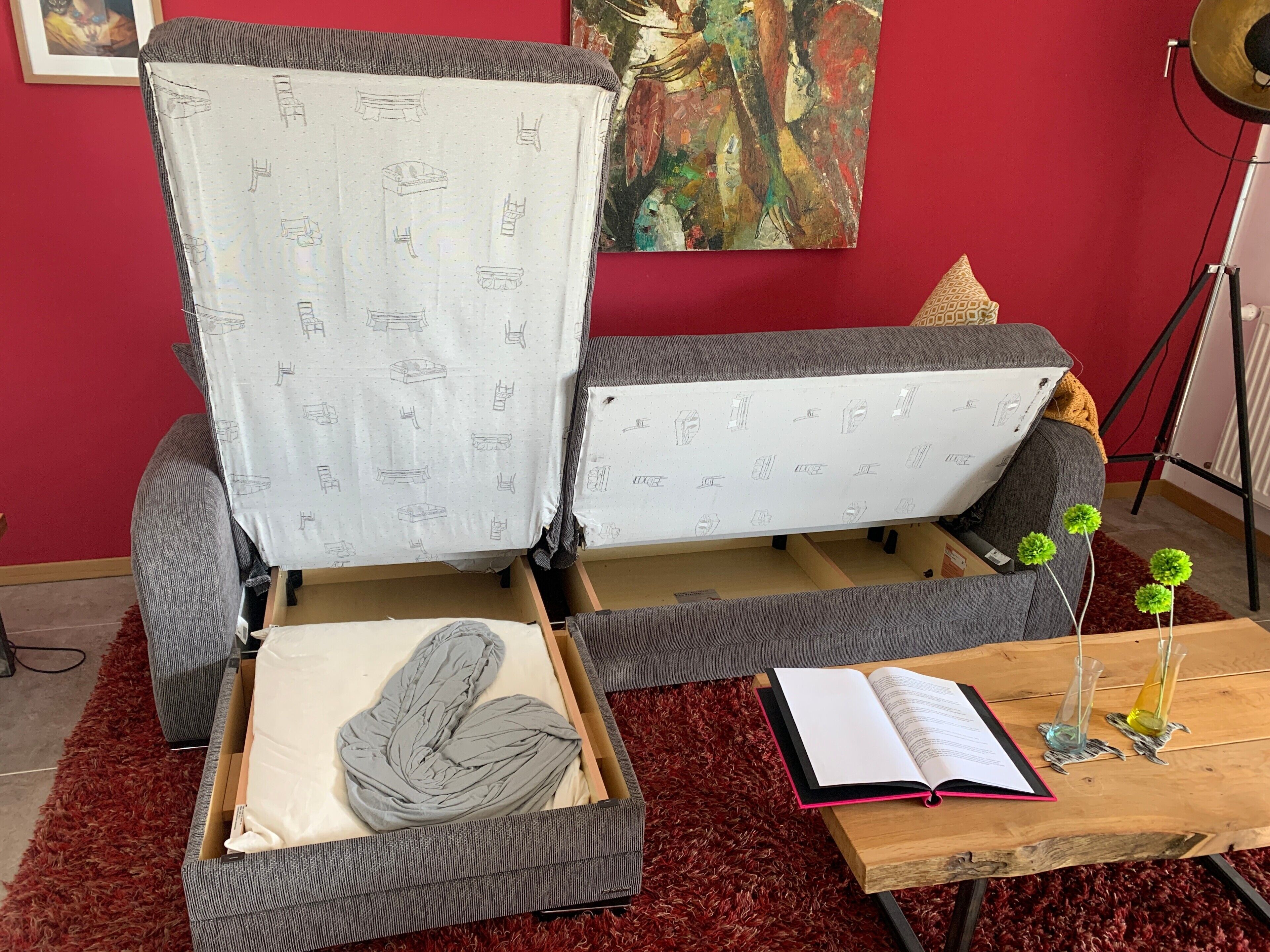 Une pièce avec un canapé gris, une moquette rouge, une table basse en bois sur laquelle se trouvent un livre et des objets décoratifs, et un tableau accroché au mur.