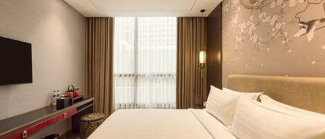 A hotel room with a large bed, a flat-screen TV mounted on the wall, a desk with a chair, and a window with curtains.