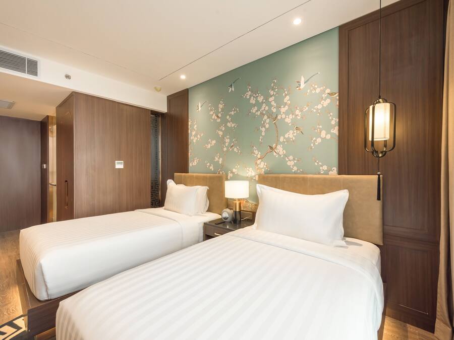 Habitació d'hotel amb dos llits, un armari de fusta, un mural a la paret, una tauleta de nit amb una làmpada i una llum penjant.