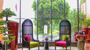 A room with two black chairs with red and green cushions, a small table with a vase, and a large window with a view of trees and buildings. - Ashley Tang Menteng Jakarta (Jakarta)