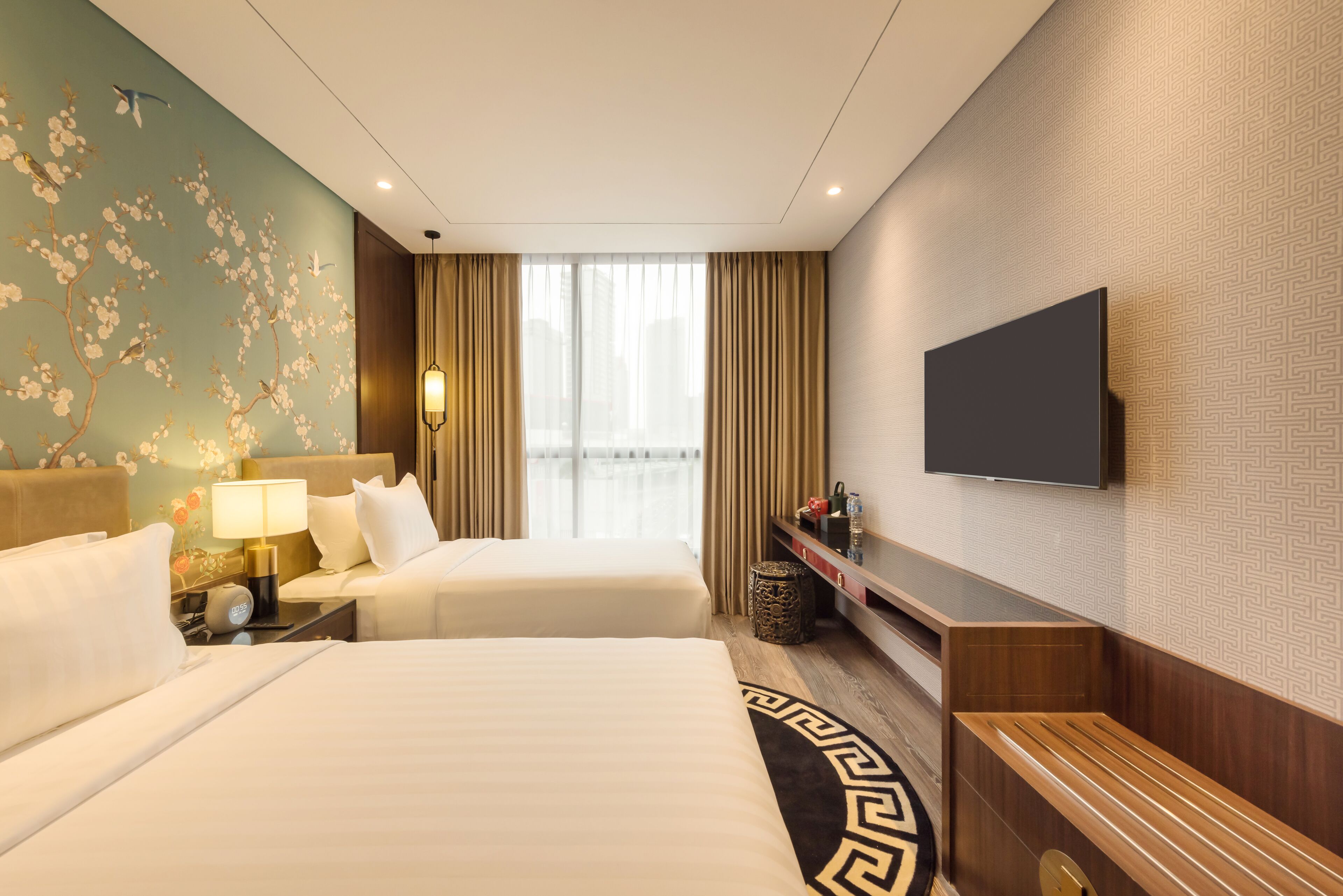 A hotel room with two beds, a TV, a desk, and a large window with curtains.