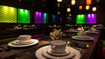 Un restaurant avec de nombreuses tables, chacune agrémentée de tasses et de soucoupes blanches, éclairées par des panneaux muraux aux couleurs vives et des lanternes suspendues.