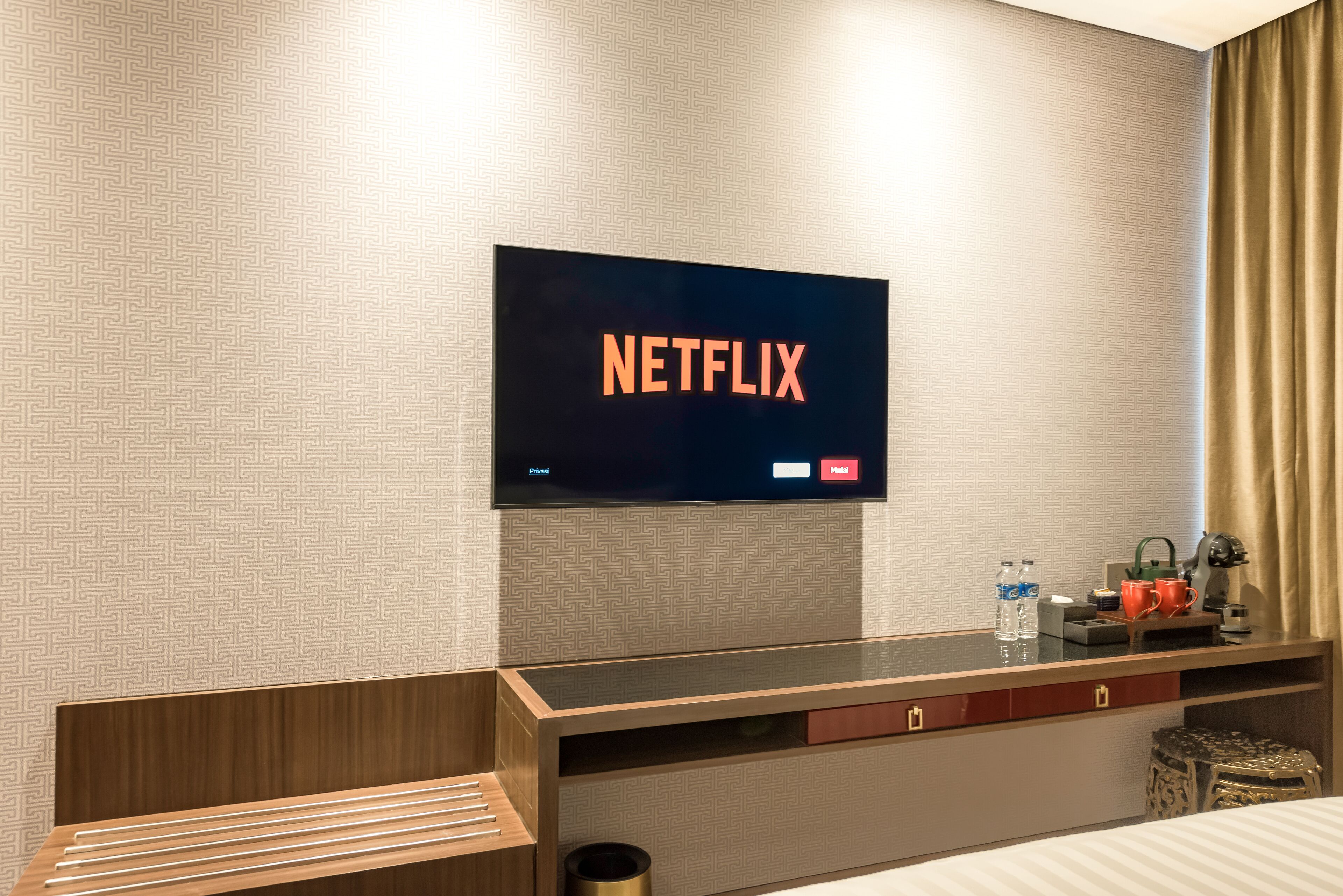 A hotel room with a flat-screen TV displaying the Netflix logo, a wooden desk with a cup, a bottle, and a small plant.