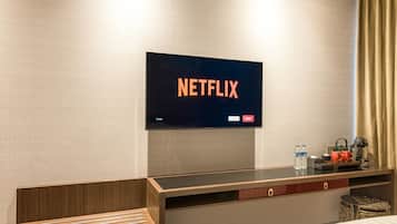 Une chambre d’hôtel équipée d’un téléviseur à écran plat affichant le logo de Netflix, d’un bureau en bois sur lequel se trouvent une tasse, une bouteille et une petite plante.