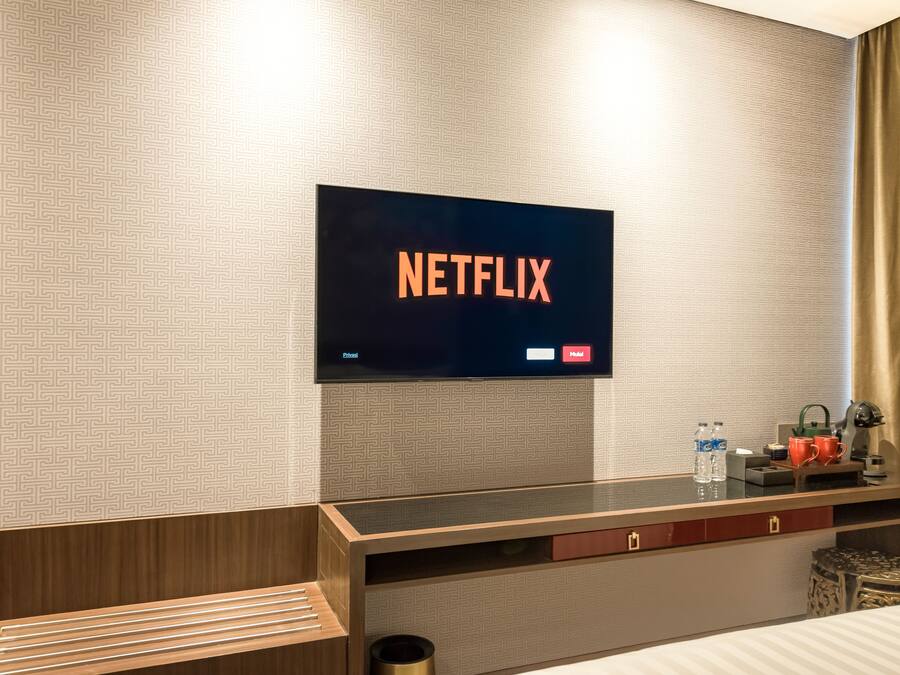Une chambre d’hôtel équipée d’un téléviseur à écran plat affichant le logo de Netflix, d’un bureau en bois sur lequel se trouvent une tasse, une bouteille et une petite plante.