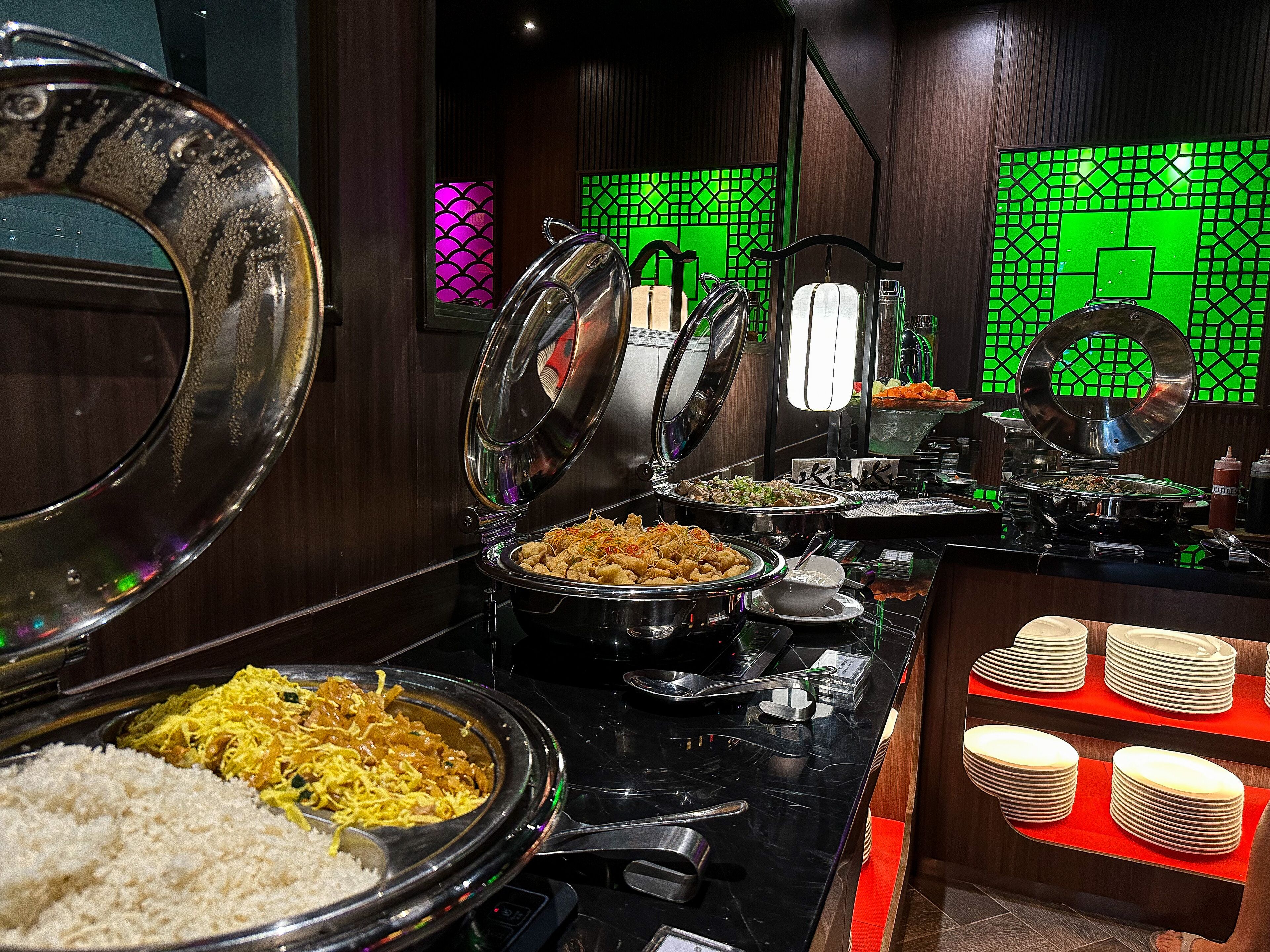 A buffet setup with various dishes, including rice, noodles, and salads, in a well-lit room with wooden paneling and green decorative panels.