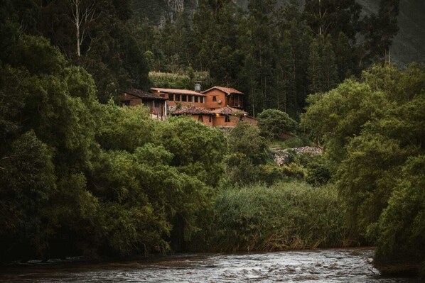 A rustic house surrounded by dense forest, with a river in the foreground.
