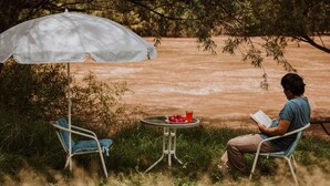 Eine Person sitzt an einem Tisch unter einem weißen Sonnenschirm und liest ein Buch. Ein Stuhl und ein Tisch mit Getränken und Obst befinden sich in der Nähe.