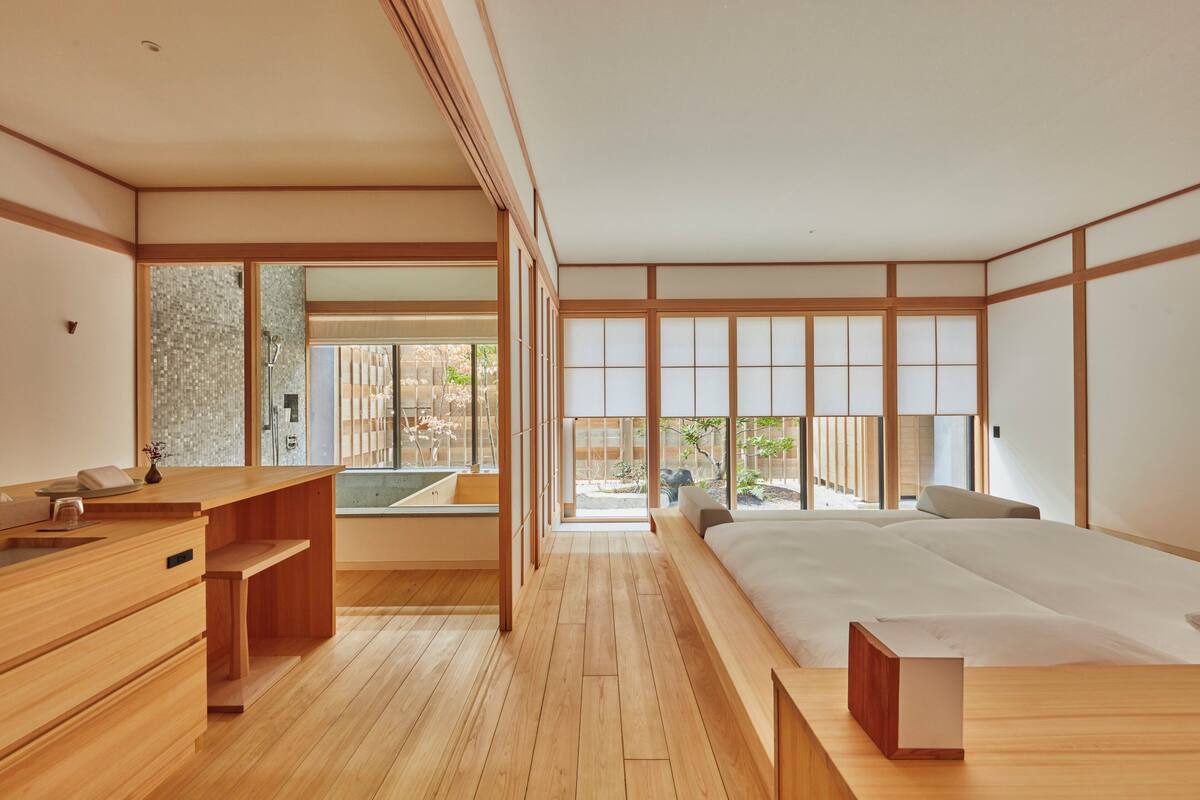 A modern Japanese-style bedroom with a wooden floor, a large bed, a wooden desk, and a view of a garden through large windows.